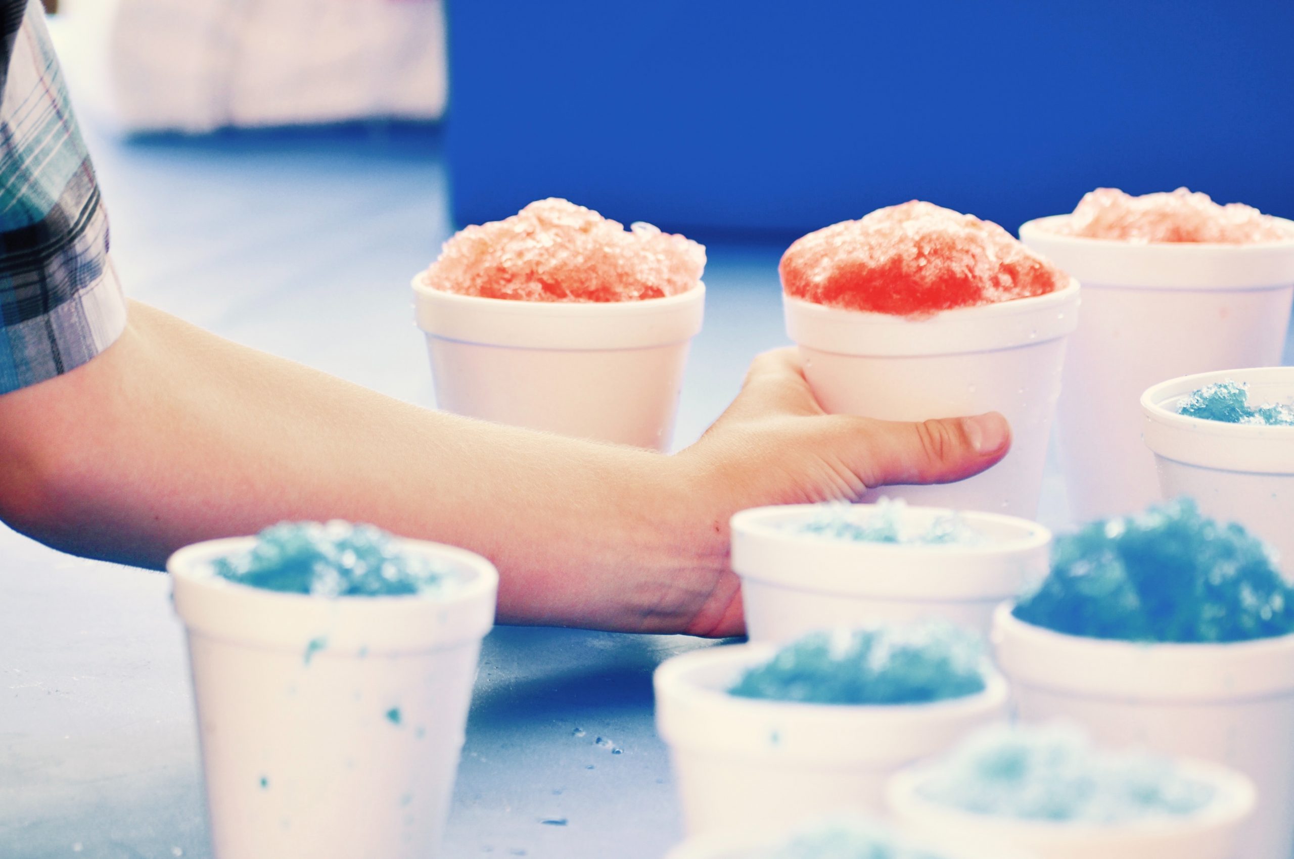 Multiple servings of Hawaiian shave ice sit on a table. A hand reaches in to grab a cherry flavored serving.